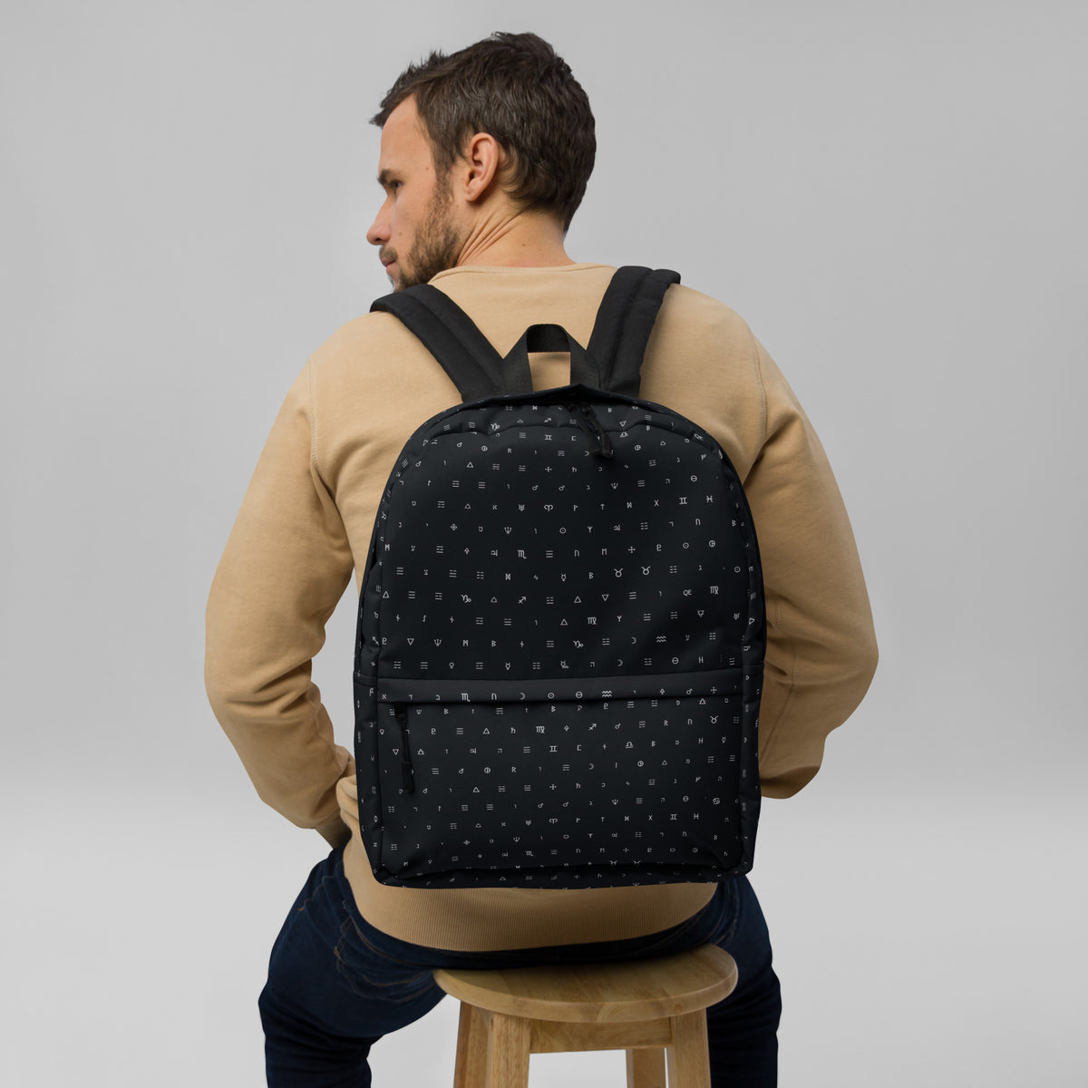 Man on a wooden stool wearing a black backpack gridded with tiny white alchemical symbols